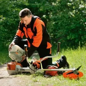 a man wearing orange brushcutter PPE (minus the helmet), kneeling down to fill the fuel tank of a STIHL petrol brushcutter