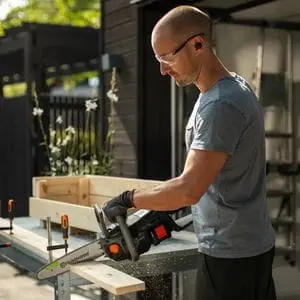 a man wearing safety glasses, using the Husqvarna Aspire™ C15X-P4A Cordless Chainsaw to cut a piece of wood on a sawhorse 