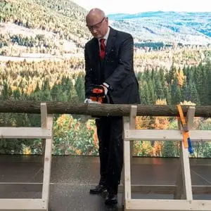 Dr Nikolas Stihl dressed in a suit, using a STIHL chainsaw to cut a thin log on a sawhorse, with an epic forest scene in the background