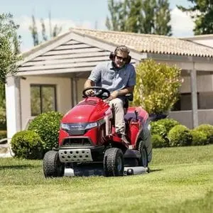 a man wearing sunglasses and ear defenders, using the Honda HF 2625 HTE Premium Petrol Lawn Tractor to mow a lawn