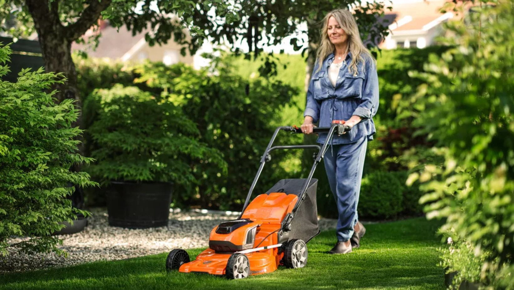 an older woman with long hair and wearing a blue outfit, mowing a suburban lawn with a Husqvarna LC 142i Lawnmower
