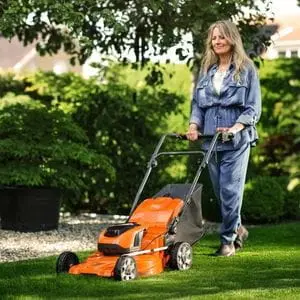 a woman with long, grey hair, using a Husqvarna LC 142iS Lawnmower to cut grass in a suburban garden