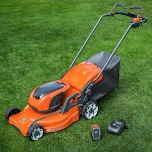 an orange cordless Husqvarna mower on a lawn, next to a battery and charger