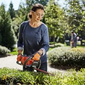 a woman wearing safety glasses, using a Husqvarna hedge cutter to trim the top of a hedge in a leafy residential neighbourhood