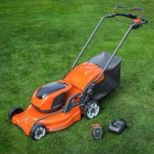 an orange Husqvarna cordless mower, next to a battery and charger, all on a lawn