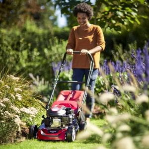 a smiling woman mowing a lawn between flowerbeds using the Mountfield SP46 Petrol Lawnmower