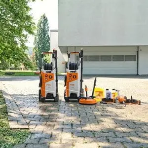 two STIHL RE 150 PLUS Electric Pressure Washers, with detergents and STIHL patio cleaner attachments, on a driveway outside a white commercial premises