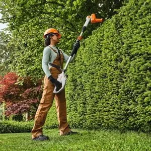 a woman wearing an orange hardhat and brown dungarees, using the STIHL HLA 40 Cordless Long-Reach Hedge Trimmer to cut the top of a tall hedge