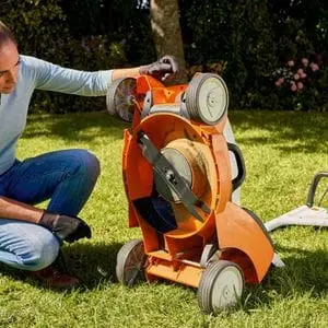 a woman inspecting the underside of an orange STIHL lawnmower in a garden