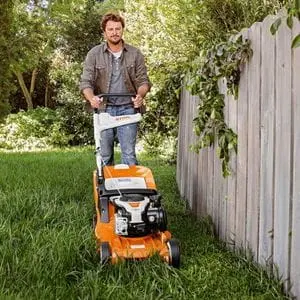 a man mowing a lawn next to a fence, using a STIHL lawnmower