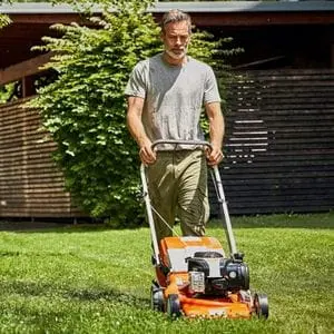 a man mowing a lawn using an orange STIHL mower, with a tree and slatted fence panels in the background