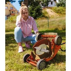 an unhappy-looking woman crouched down next to an upturned electric lawn mower