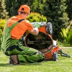a technician dressed in an orange and green uniform, inspecting the underside of a lawnmower