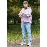 a man wearing glasses and a pink hooded top, stood in a park, looking at his watch