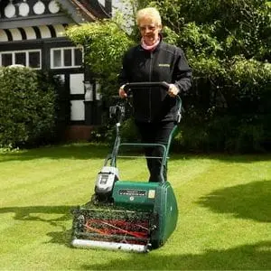 an elderly woman wearing glasses, using the Allett Cambridge 43 Cordless Cylinder Mower to cut a lawn outside an attractive period home