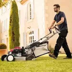a man wearing protective glasses, using the Honda HRD 536 QX Petrol Lawn Mower to mow a lawn outside a stately home