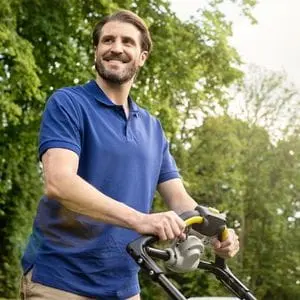a smiling, bearded man standing in a garden, holding a lawnmower's handle