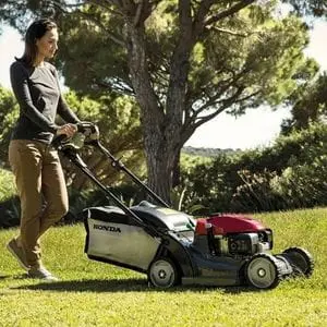 a woman cutting grass in front of trees and hedging, using the Honda HRX 476 HY Petrol Lawn Mower