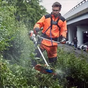 a man wearing orange, high-visibility PPE, using the Husqvarna 535iRXT Cordless Brushcutter to cut back vegetation on a roadside verge 