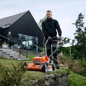 a man using the Husqvarna LB 553iV Cordless Mower to cut grass on a steep and narrow strip of grass outside a Scandinavian-style house