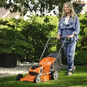 a woman with long grey hair, mowing a suburban lawn using the Husqvarna LC 142i Cordless Lawn Mower