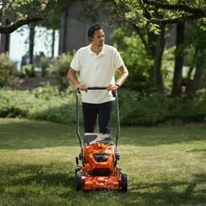 a man mowing a lawn in front of some trees, using the Husqvarna LC 251iS Cordless Mower