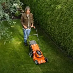 a woman mowing a lawn, between a tree and hedge, using the Husqvarna LC137i Cordless Lawnmower