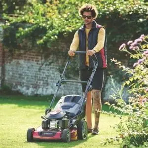 a man wearing sunglasses, using the Mountfield HW531 PD Petrol Lawn Mower to mow a lawn near a wall and shrubs