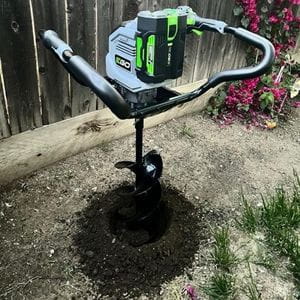 the EGO PHA7404E Cordless Earth and Ice Auger Power Head and auger bit stood in a hole in the ground, next to a fence