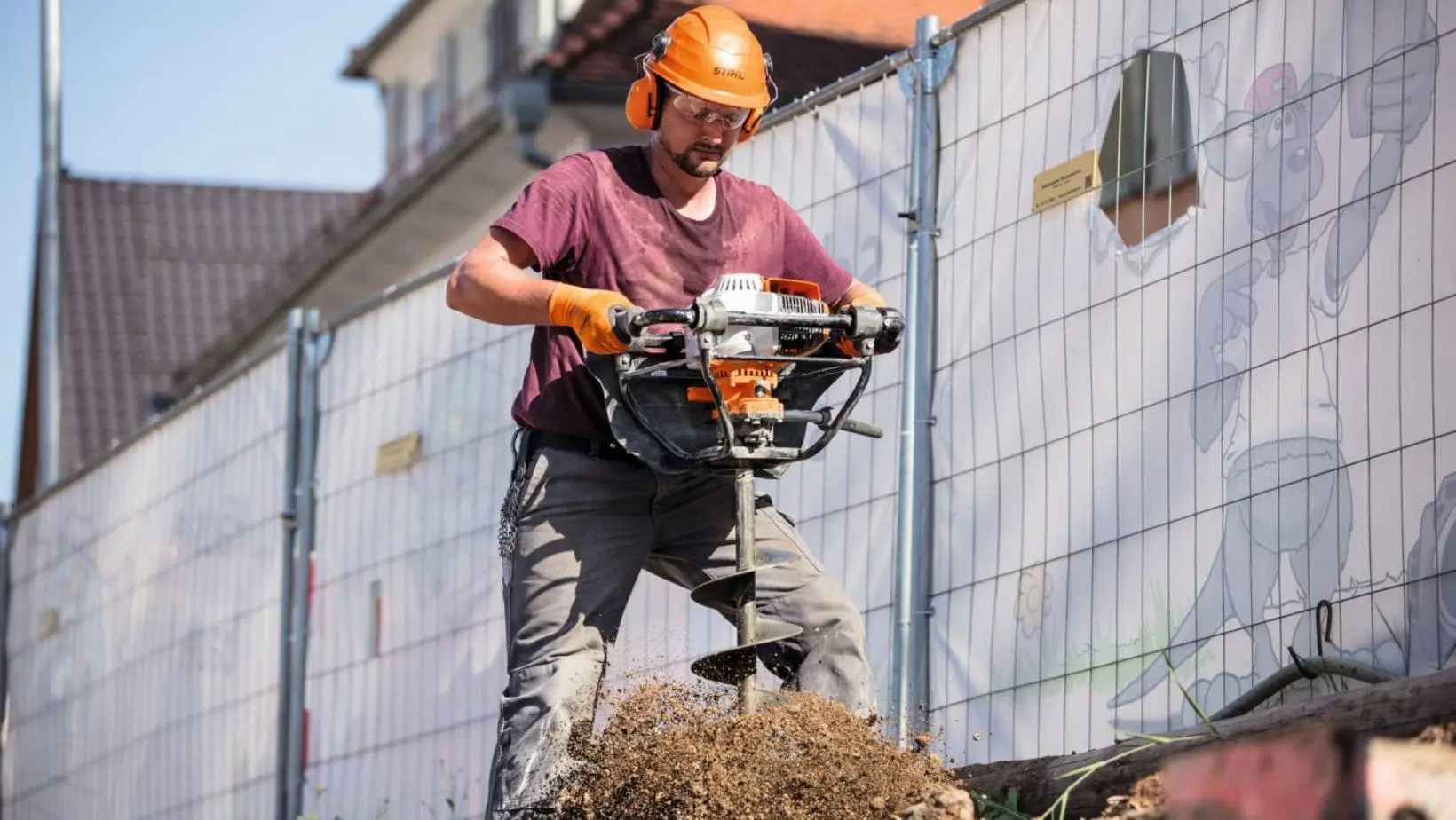 a man wearing a hardhat and safety glasses, using the STIHL BT 131 Petrol Earth Auger to drill a hole in the ground next to a newbuild housing development