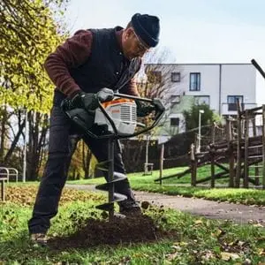 a man wearing a beanie hat and safety glasses, crouched over the STIHL BTA 140 Cordless Earth Auger as it drills a hole in the ground, near a children's play area