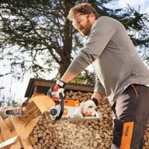 a man using the STIHL MSA 70 Cordless Chainsaw to cut a log on a sawhorse, with a large pile of logs in the background