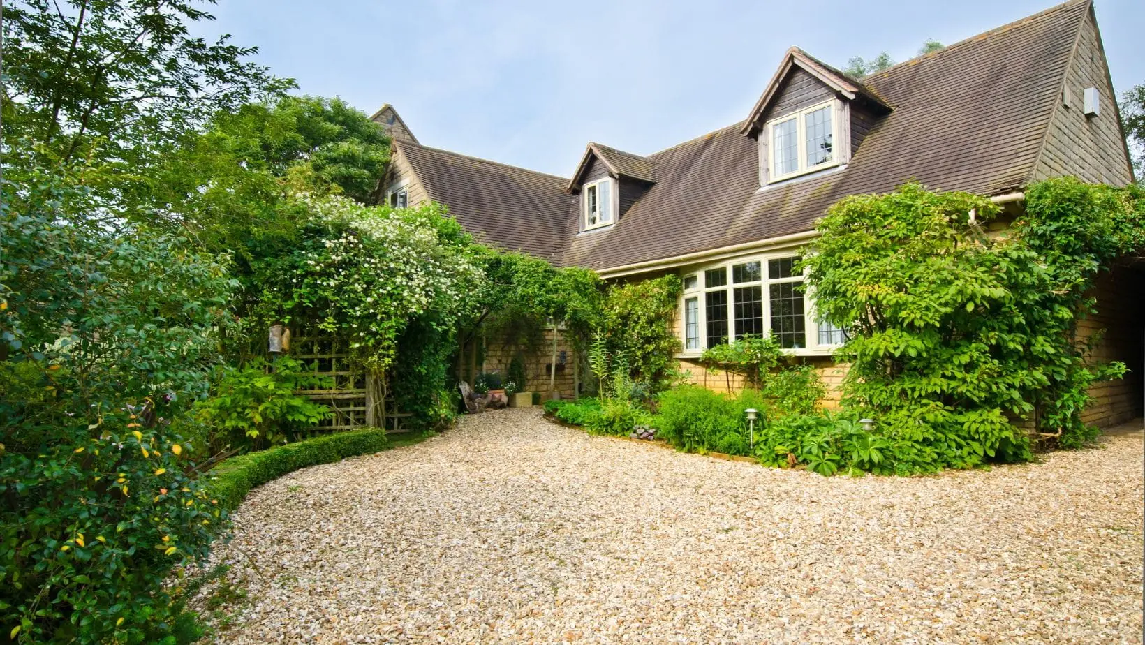 Gravel driveway leading to a traditional house surrounded by lush green shrubs, trees and garden borders.