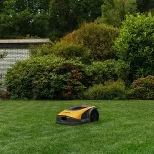 a STIGA Vista A 10V Robotic Mower cutting a lawn, with trees, shrubs and an outbuilding in the background