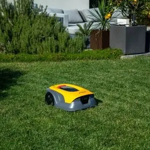 a STIGA Vista A 6V Robotic Mower on a lawn, with shrubs, potted plants and patio furniture in the background