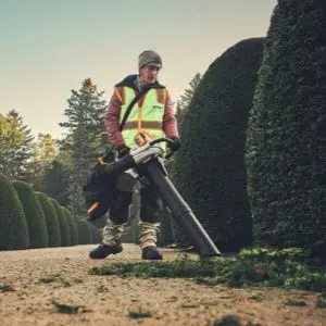 Groundskeeper using a cordless leaf vacuum shredder to clear fallen leaves along a gravel path beside neatly trimmed hedges.
