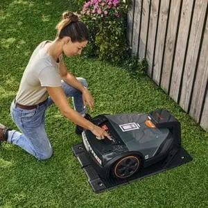 a woman knelt on one knee on a lawn, programming a STIHL robotic mower sat in its charging station