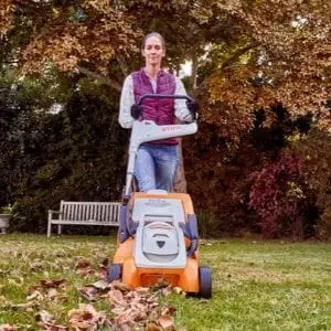 a woman wearing a gilet, using a STIHL mower to cut a lawn covered in autumn leaves
