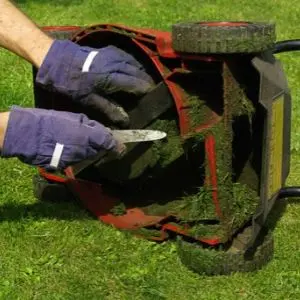 someone wearing gloves, using a small knife to remove wet grass clippings from the underside of a lawnmower